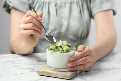 Woman eating tasty yogurt with fresh kiwi at white marble table, closeup