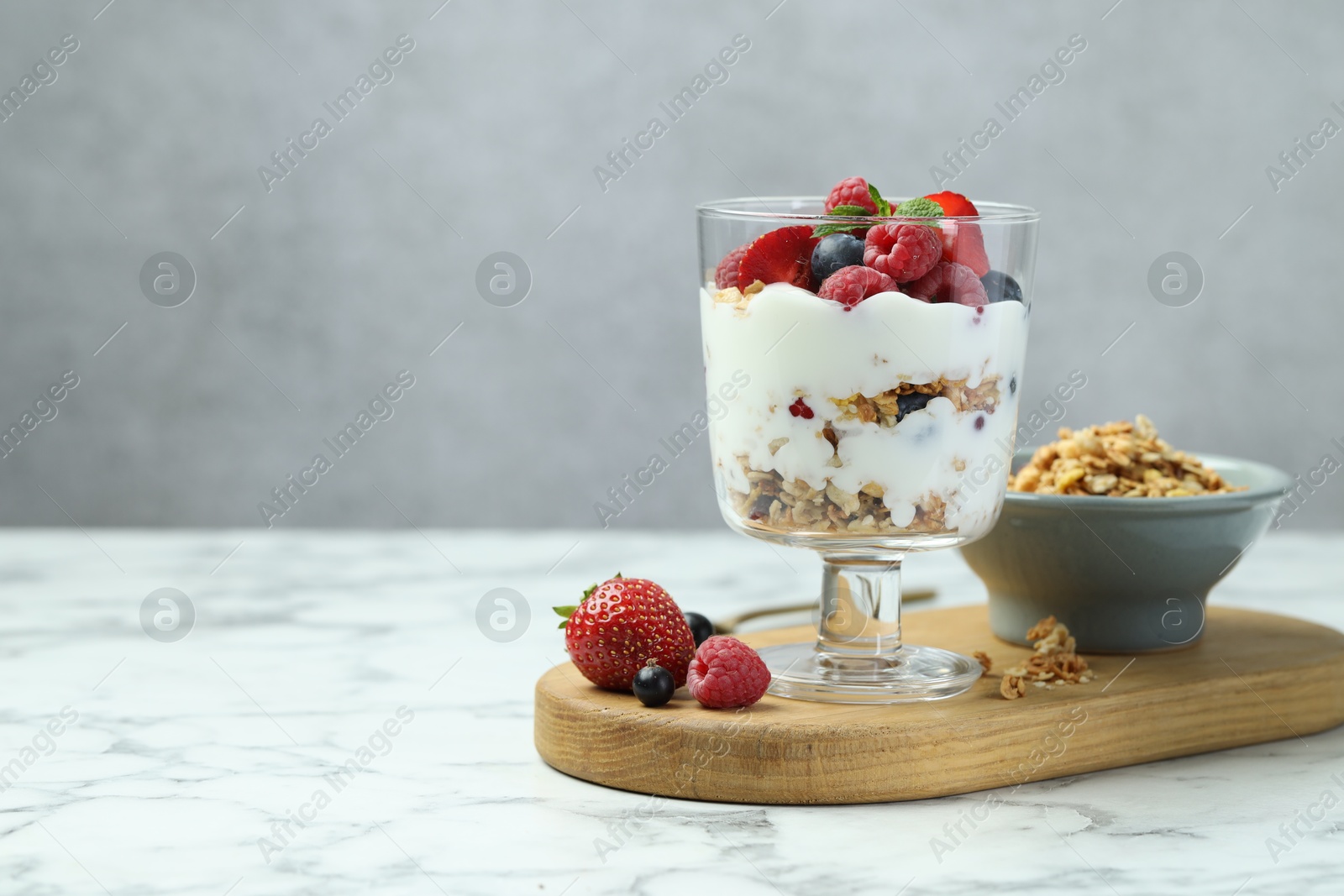 Photo of Tasty yogurt with fresh berries and granola in glass dessert bowl on white marble table, space for text