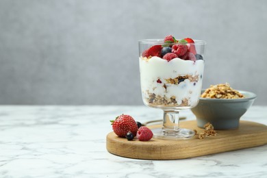 Tasty yogurt with fresh berries and granola in glass dessert bowl on white marble table, space for text