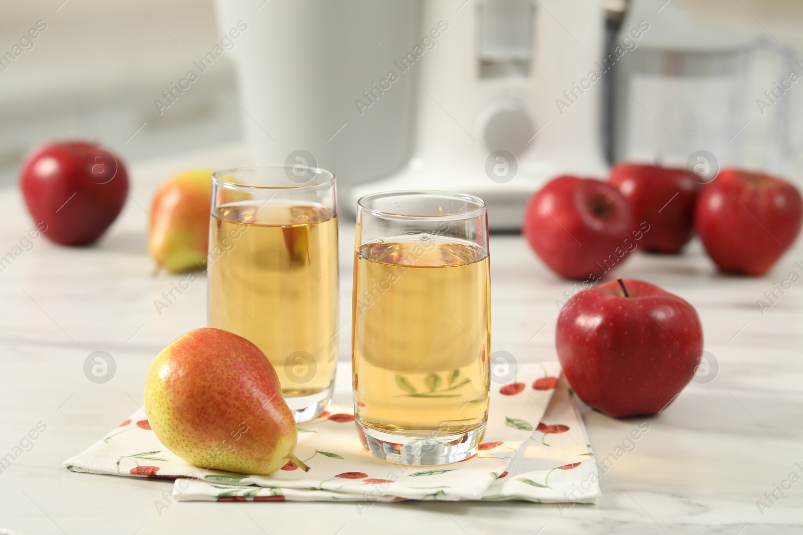 Photo of Glasses of fresh juice, pears, apples and modern juicer on white marble table in kitchen, selective focus