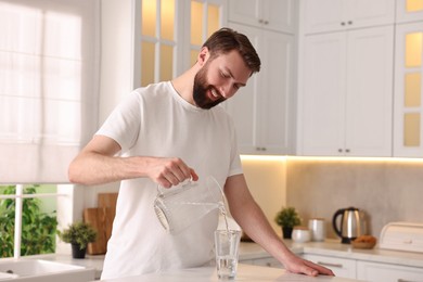 Photo of Happy young man pouring water into glass in kitchen at morning