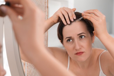 Photo of Hair loss problem. Woman applying serum onto hairline near mirror indoors