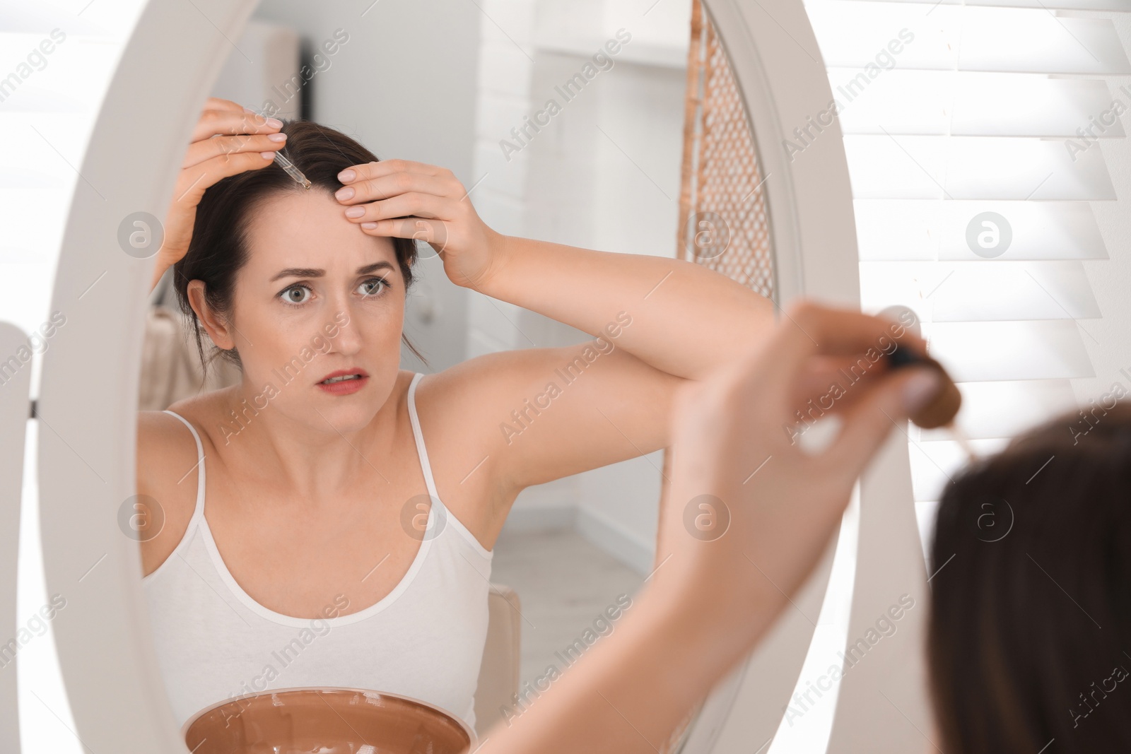 Photo of Hair loss problem. Woman applying serum onto hairline near mirror at home