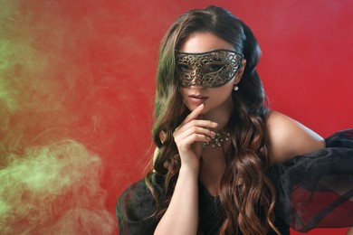 Photo of Beautiful woman wearing carnival mask on red background in color lights and smoke. Space for text