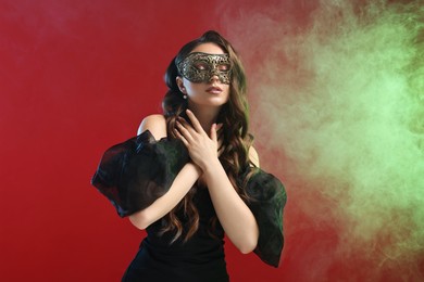 Photo of Beautiful woman wearing carnival mask on red background in color lights and smoke