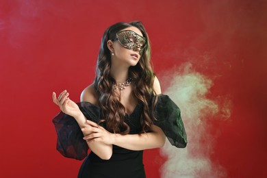 Photo of Beautiful woman wearing carnival mask on red background in color lights and smoke