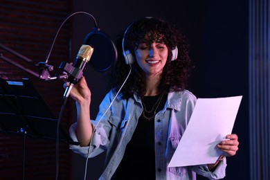 Photo of Woman with headphones singing into microphone in professional record studio