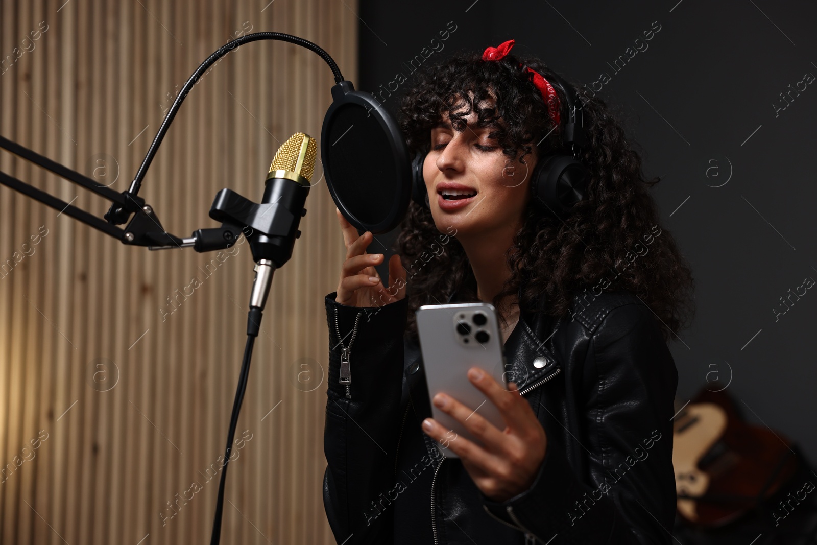 Photo of Woman wearing headphones with smartphone singing into microphone in professional record studio