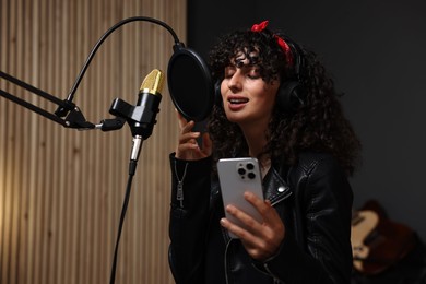 Photo of Woman wearing headphones with smartphone singing into microphone in professional record studio