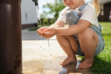 Photo of Water scarcity. Little girl drawing water with hands from tap outdoors, closeup