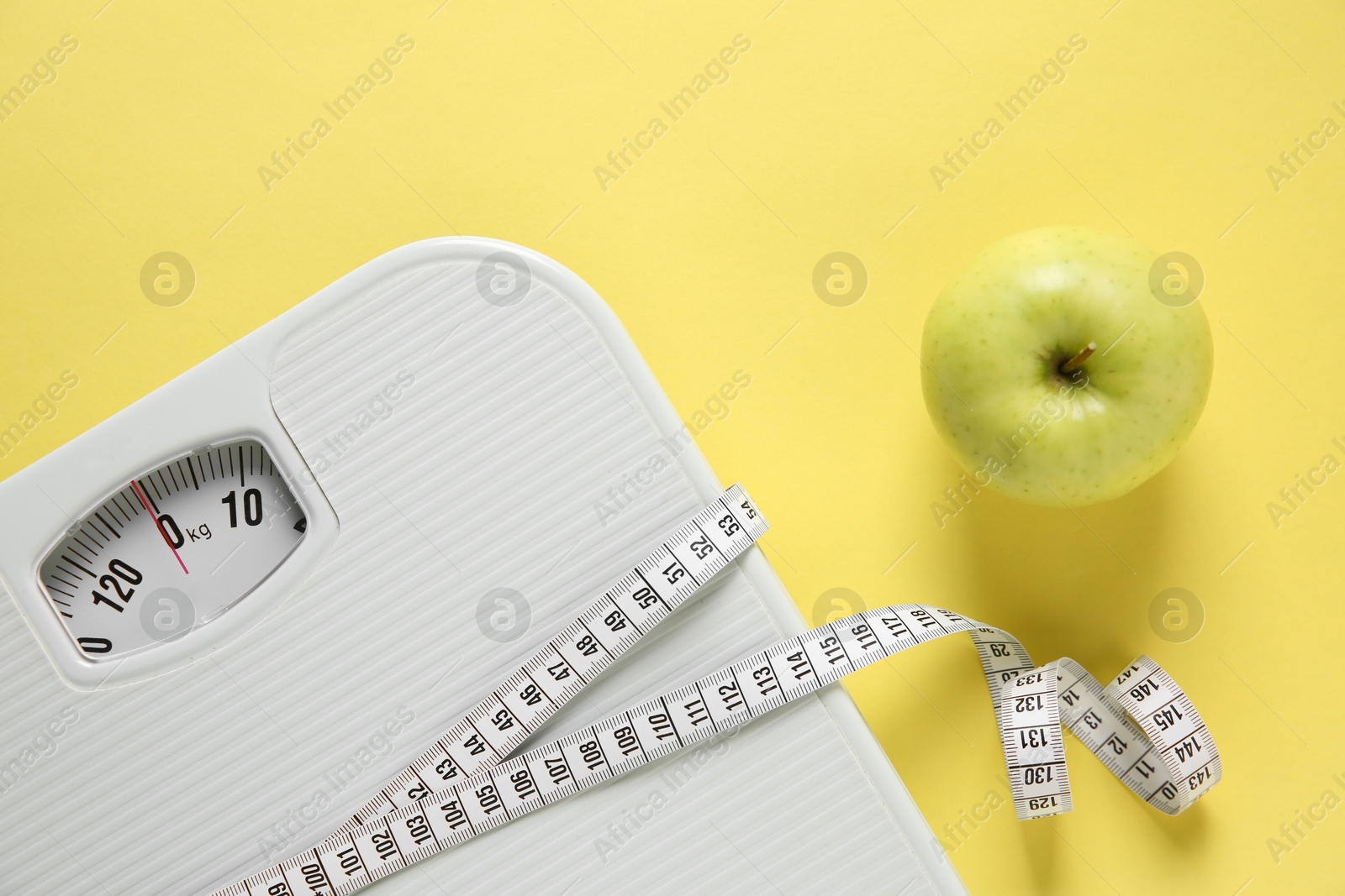 Photo of Scale tied with measuring tape and fresh apple on yellow background, flat lay