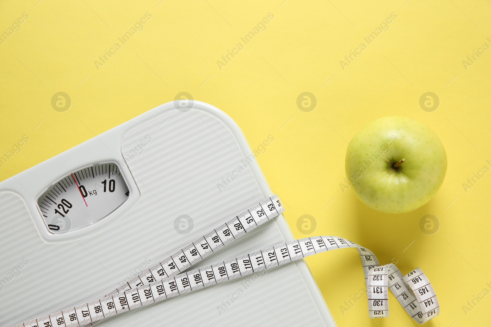 Photo of Scale tied with measuring tape and fresh apple on yellow background, flat lay