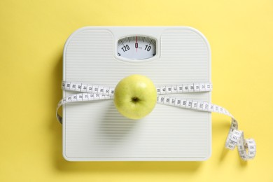 Photo of Scale tied with measuring tape and fresh apple on yellow background, top view