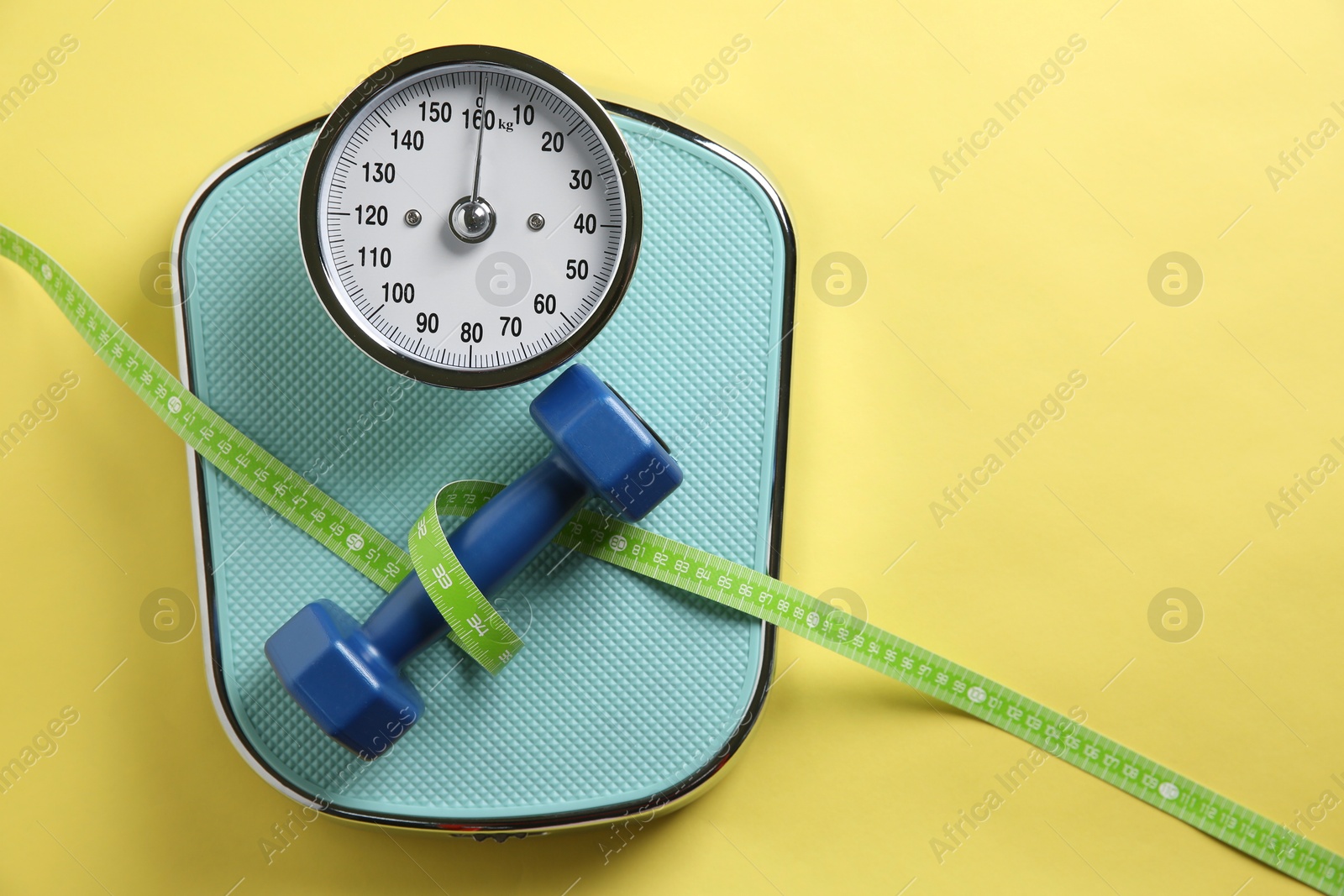 Photo of Scale, measuring tape and dumbbells on yellow background, top view