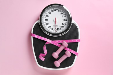 Photo of Scale tied with measuring tape and dumbbells on pink background, top view