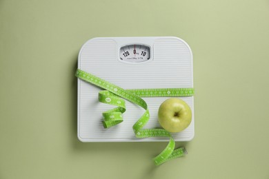 Scale tied with measuring tape and fresh apple on olive background, top view