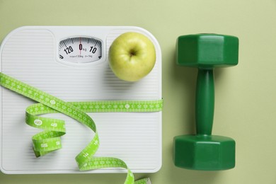 Photo of Scale, measuring tape, dumbbells and fresh apple on olive background, flat lay