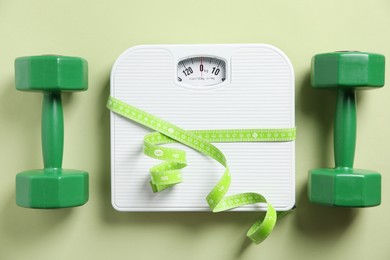 Photo of Scale tied with measuring tape and dumbbells on olive background, flat lay