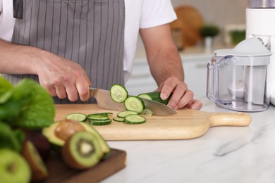 Photo of Man cutting cucumber indoors, closeup. Juicer and fresh products on white marble table