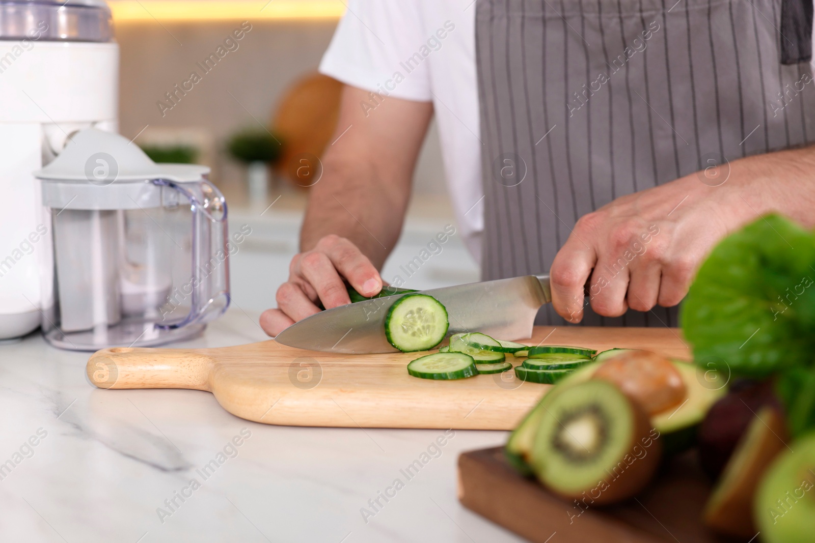 Photo of Man cutting cucumber indoors, closeup. Juicer and fresh products on white marble table