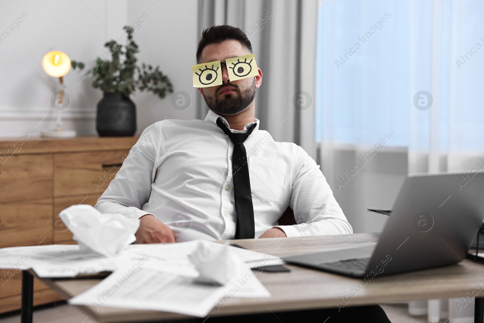 Photo of Man with fake eyes painted on sticky notes at workplace in office. Overwhelmed by work