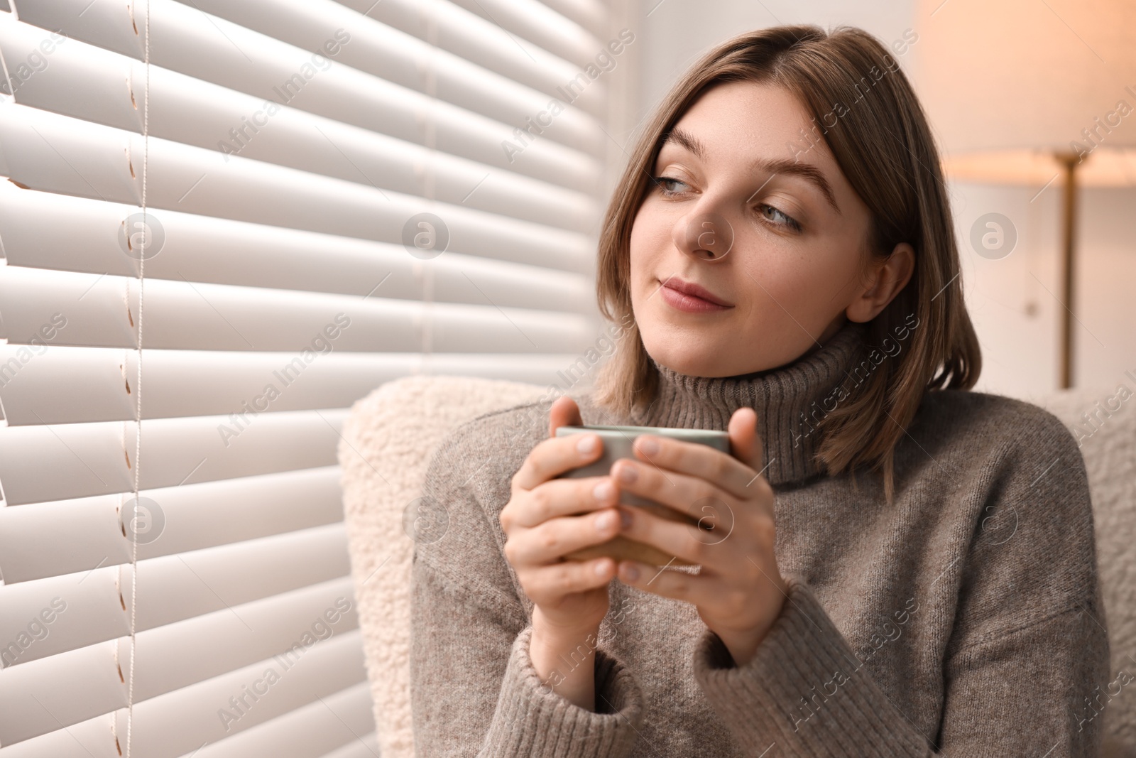 Photo of Woman with cup of drink sitting on armchair near window blinds indoors