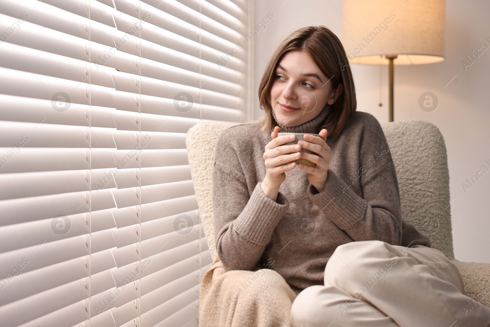 Photo of Woman with cup of drink sitting on armchair near window blinds indoors