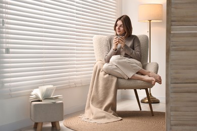 Photo of Woman with cup of drink sitting on armchair near window blinds indoors