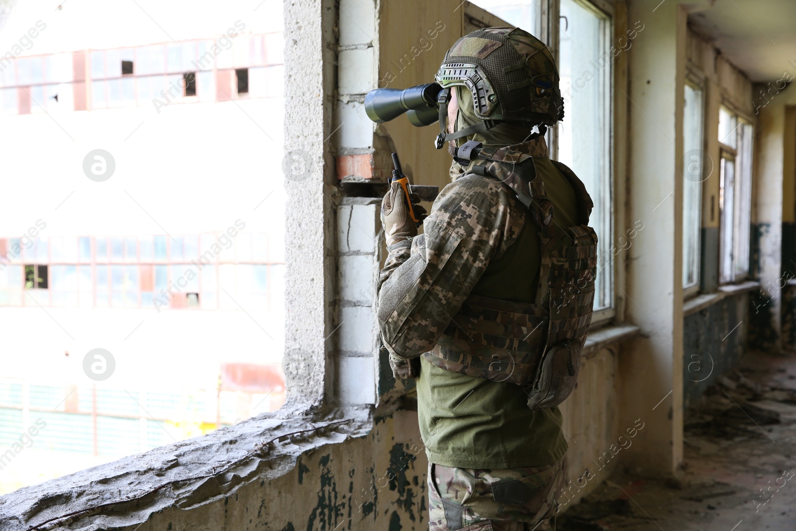 Photo of Military mission. Soldier in uniform with binoculars inside abandoned building, back view. Space for text