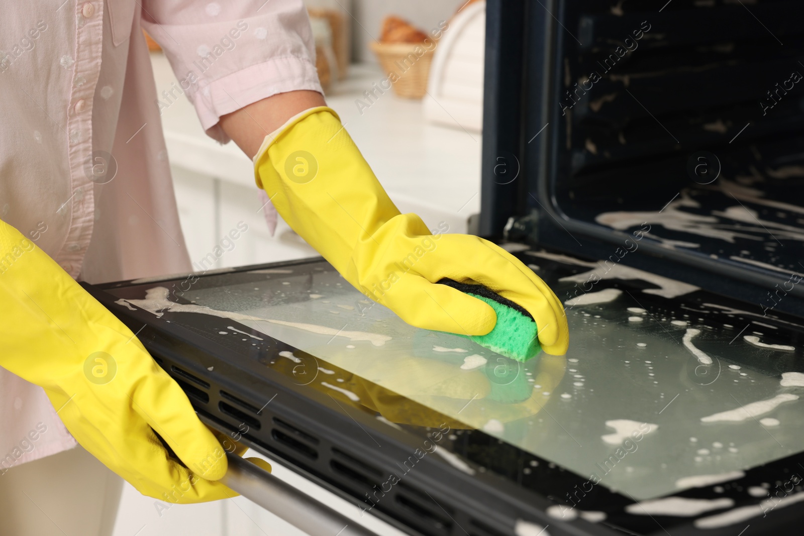 Photo of Woman cleaning oven door with sponge in kitchen, closeup