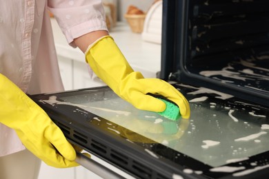 Photo of Woman cleaning oven door with sponge in kitchen, closeup