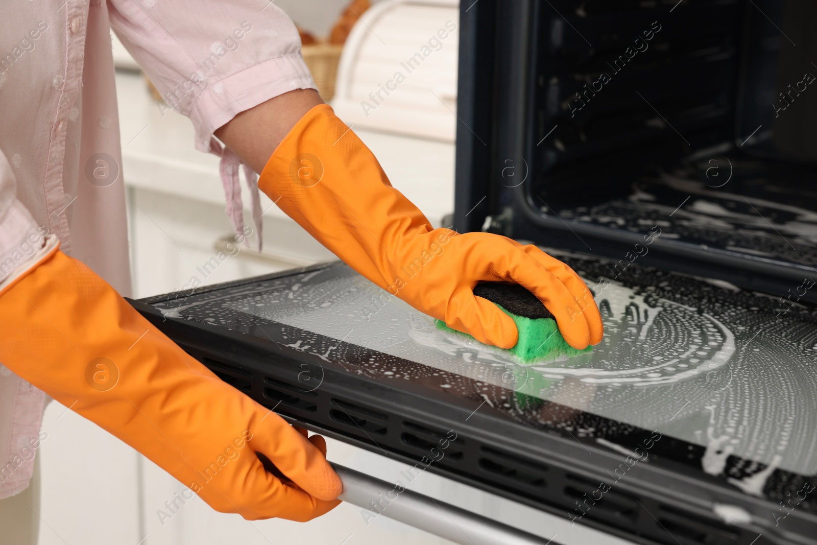 Photo of Woman cleaning oven door with sponge in kitchen, closeup