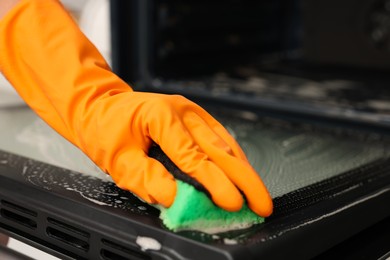 Photo of Woman cleaning oven door with sponge in kitchen, closeup