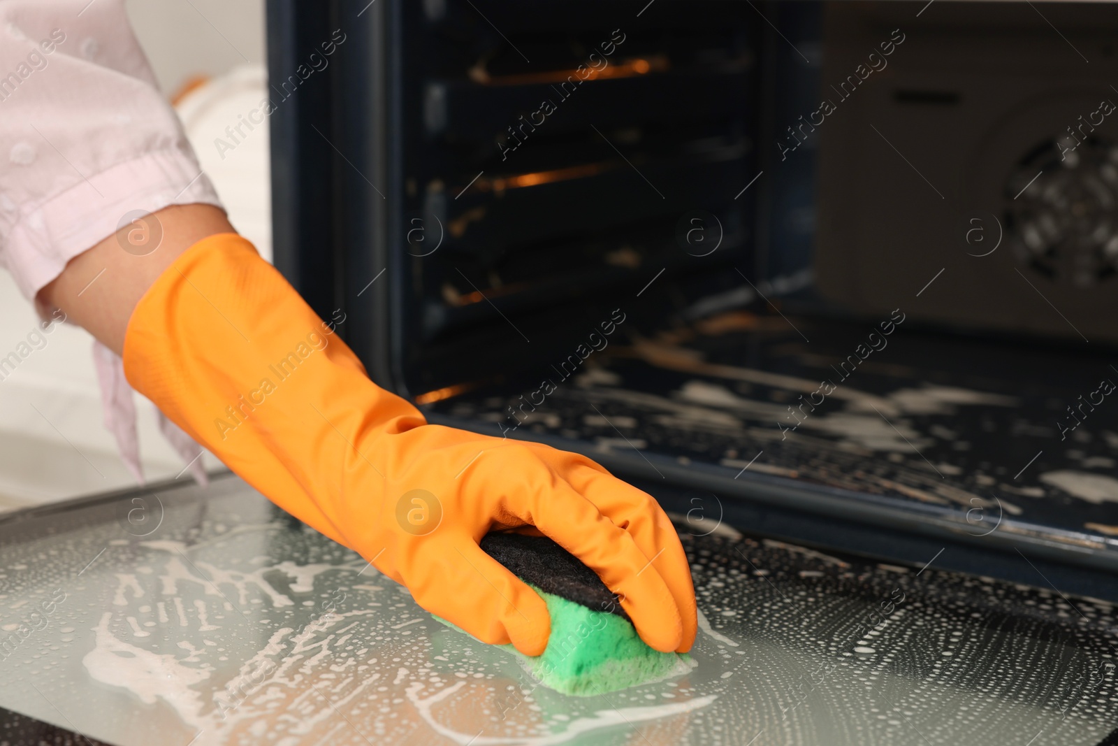 Photo of Woman cleaning oven door with sponge in kitchen, closeup