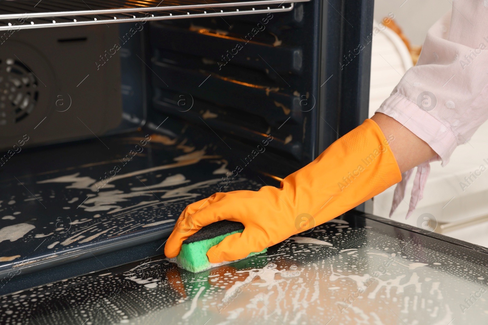 Photo of Woman cleaning oven door with sponge in kitchen, closeup