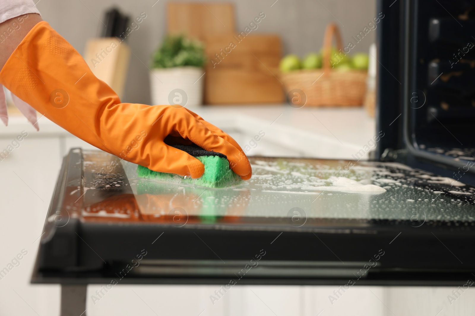 Photo of Woman cleaning oven door with sponge in kitchen, closeup