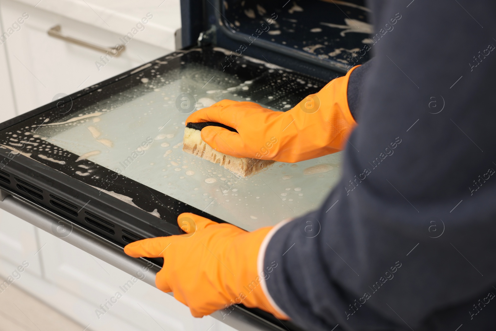 Photo of Woman cleaning oven door with sponge in kitchen, closeup