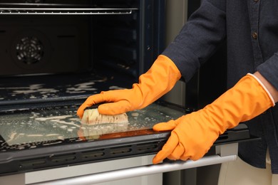 Photo of Woman cleaning oven door with sponge in kitchen, closeup