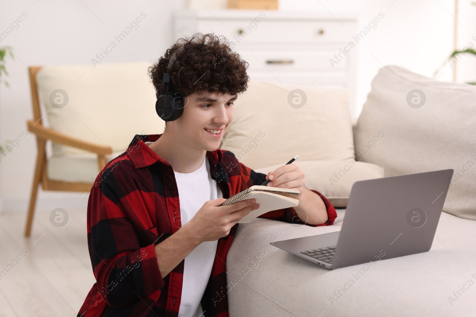 Photo of Teenager in headphones taking notes while working with laptop at home. Remote job