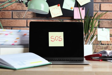 Photo of Sticky note with word SOS, laptop and stationery on wooden table at workplace