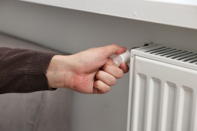 Photo of Man adjusting temperature of heating radiator indoors, closeup