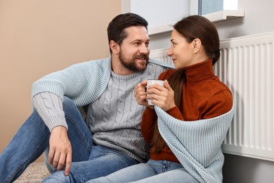 Photo of Happy couple with cup of hot drink near heating radiator at home