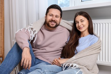 Photo of Happy couple with blanket warming near heating radiator at home
