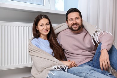 Photo of Happy couple with blanket warming near heating radiator at home