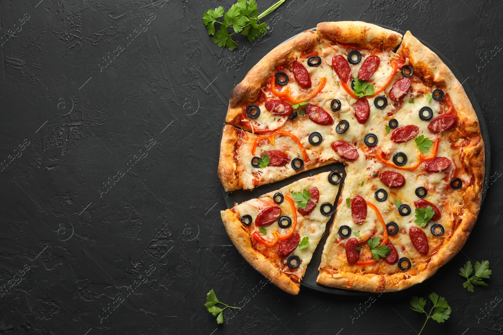 Photo of Tasty pizza with dry smoked sausages, olives, pepper and parsley on black table, top view. Space for text