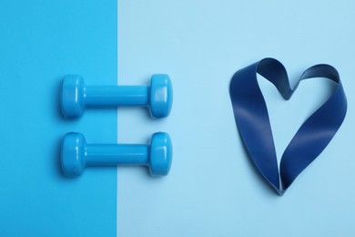 Photo of Two dumbbells and fitness elastic band in shape of heart on light blue background, flat lay. Lovely workout