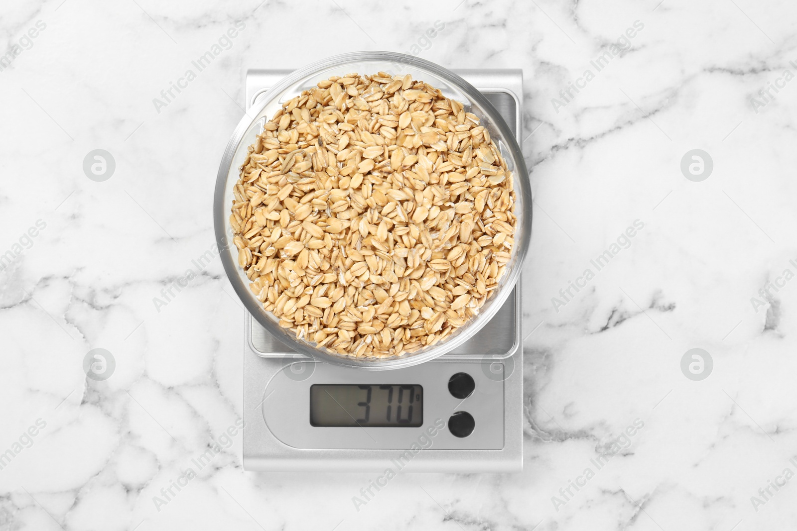 Photo of Kitchen scale with bowl of dry oats on white marble table, top view