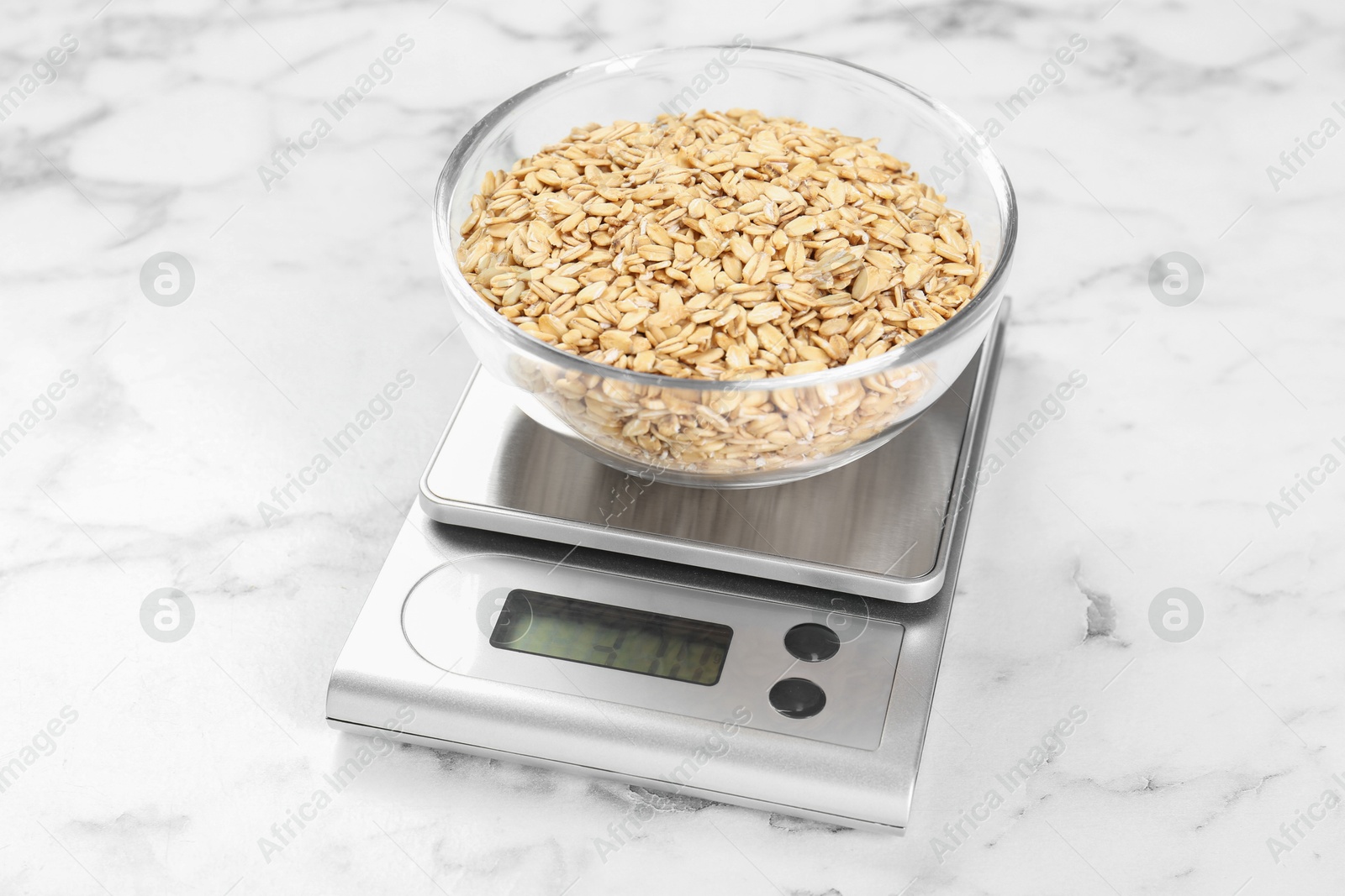 Photo of Kitchen scale with bowl of dry oats on white marble table, closeup