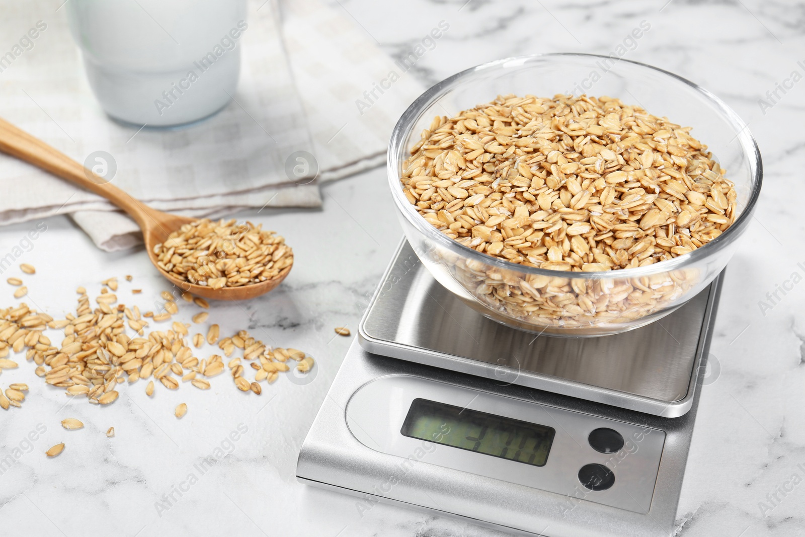 Photo of Kitchen scale with bowl of dry oats and spoon on white marble table, closeup