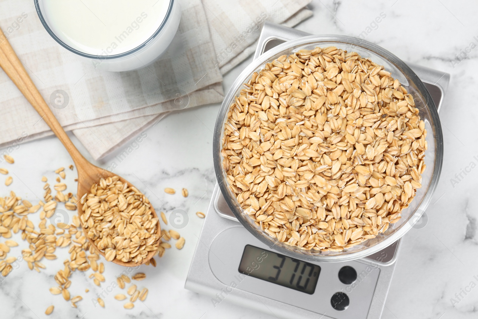 Photo of Kitchen scale with bowl of dry oats, milk and spoon on white marble table, flat lay